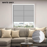 iving room with a window featuring white and gray blinds, a sofa with beige pillows, and a small table with a decorative vase.